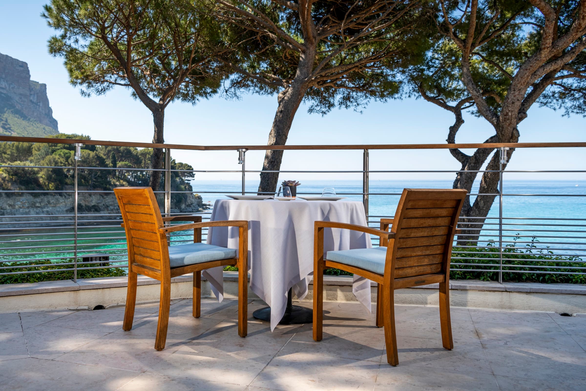 An empty round table for two on the marble cliff terrace, two oak chairs in the foreground, umbrella pines framing the view across the bay to the red-grey cliffs of Cap Canaille.
