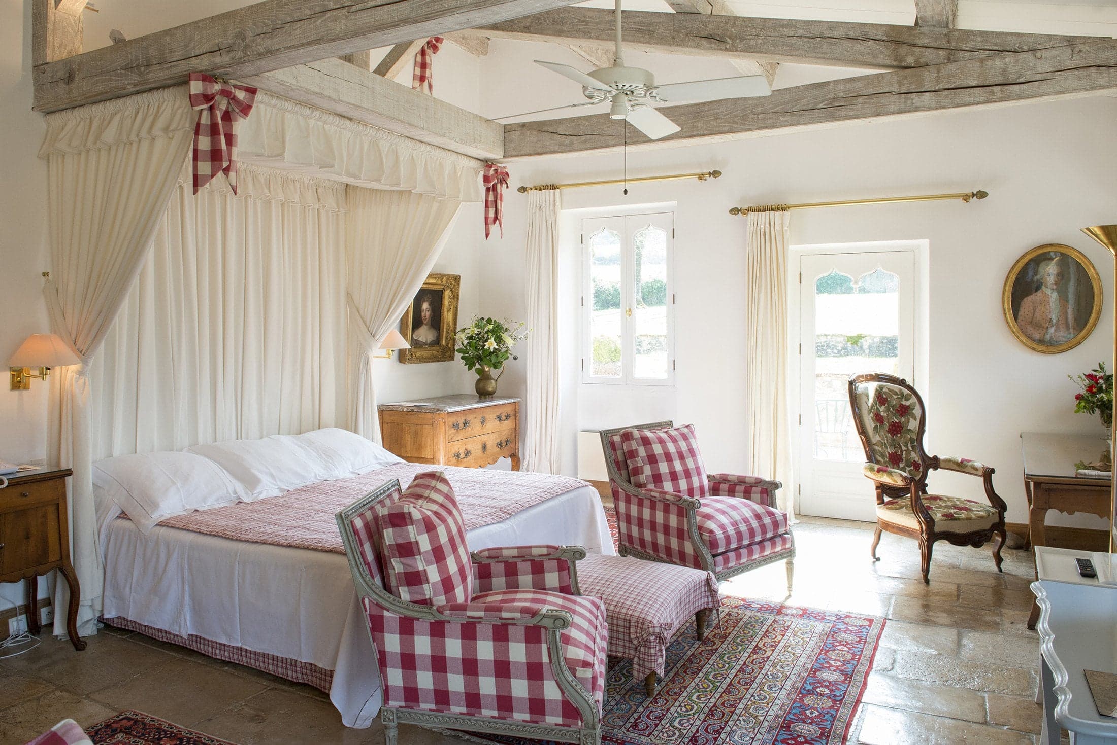 A four-poster suite under exposed beams, white linen and gingham at midday, French doors open to the terrace.