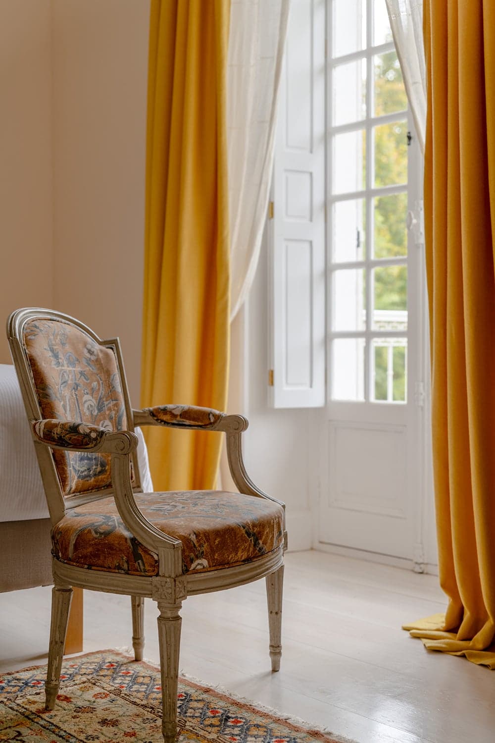 A Louis XVI armchair beside an open French window, mustard curtains drawn back to morning light.