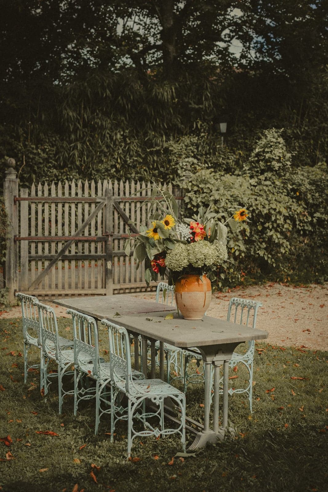 A long outdoor table at La Ferme aux Grives, sunflowers in a clay pot and pale cast-iron chairs against a picket gate.