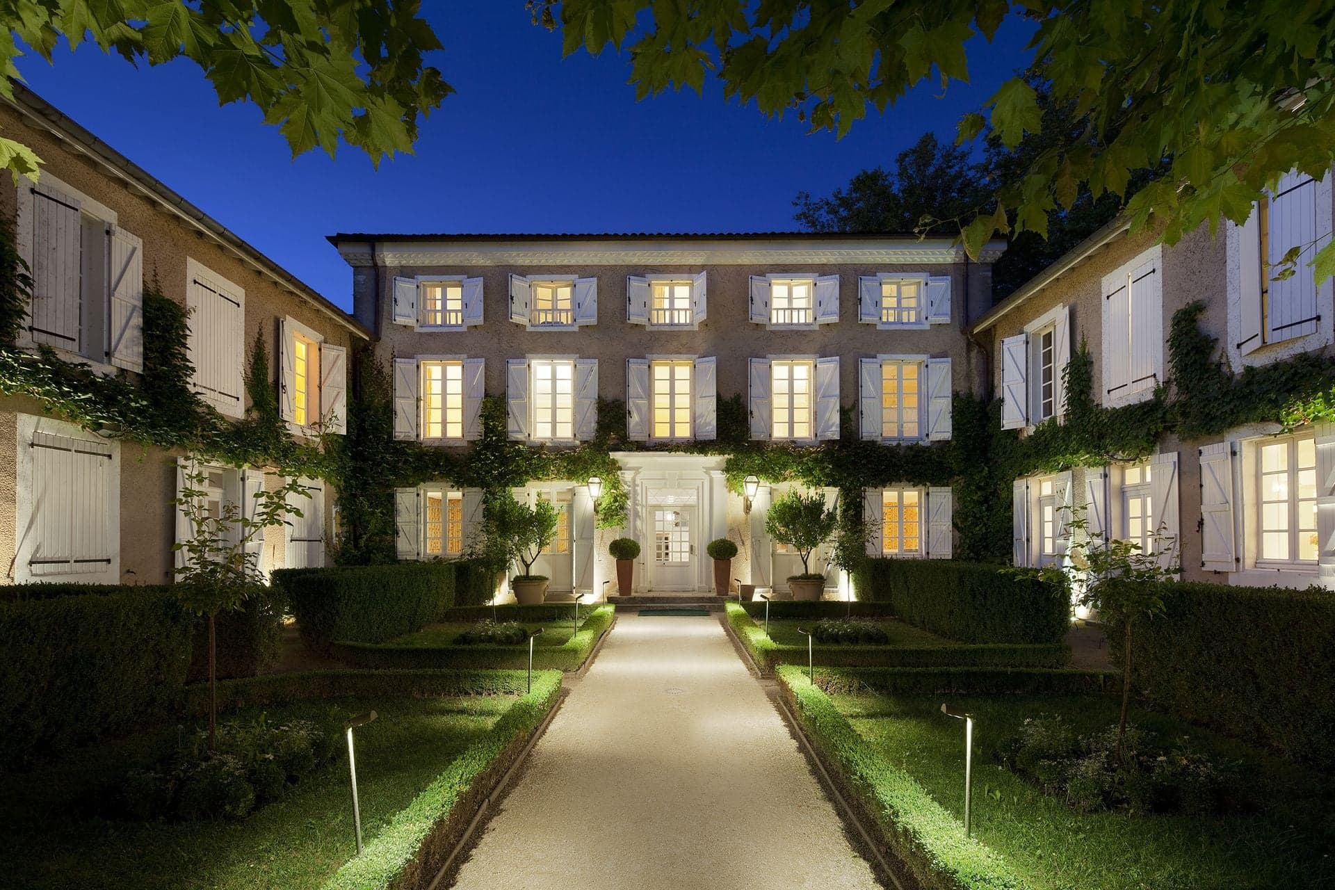 The Maison Rose at dusk, a hedged path lit between low parterres leading to its open door.