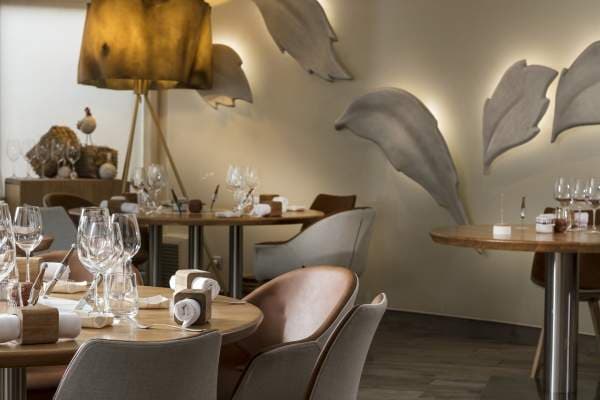 The Auberge du Vieux Puits dining room — round tables on a polished floor, chestnut leaf sculptures lit from behind on the back wall, a warm ridged lampshade over the centre table.
