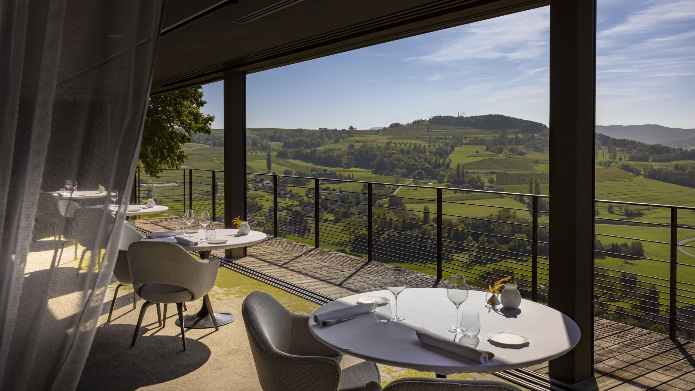 The Les Morainières dining room — three pale tables on a polished floor, a wall of plate glass opening onto rolling vineyards and the wooded ridge above, the Chapelle Saint Romain visible on a far rise.