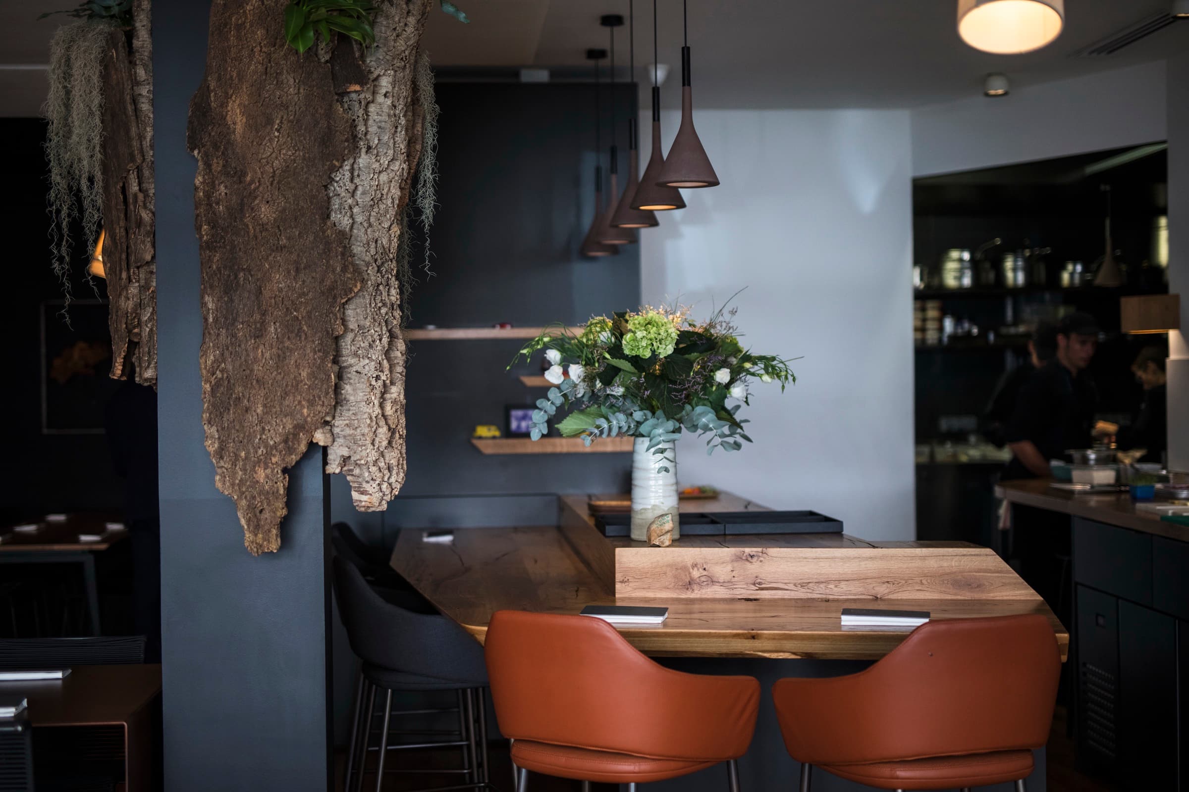 The AM dining room — a single oak counter table set with leather chairs, a hanging panel of cork bark with trailing moss, dark walls, a row of bronze pendant lights, and the open kitchen visible at the back.