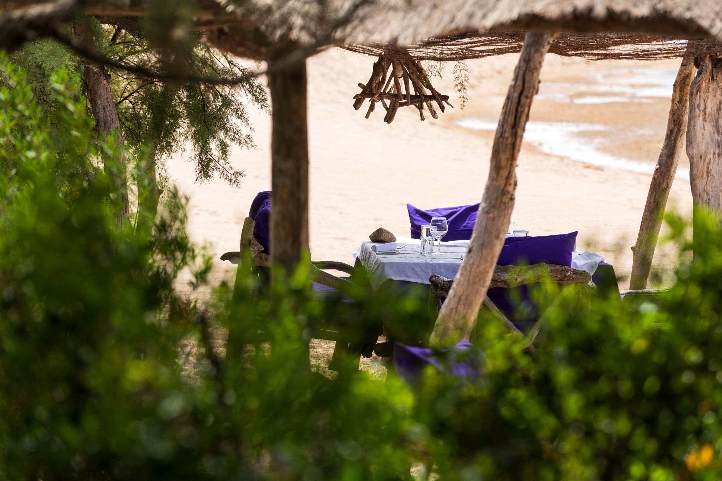 A table for two laid at La Plage, glassware on white linen, the sand and incoming surf glimpsed through driftwood and pine.