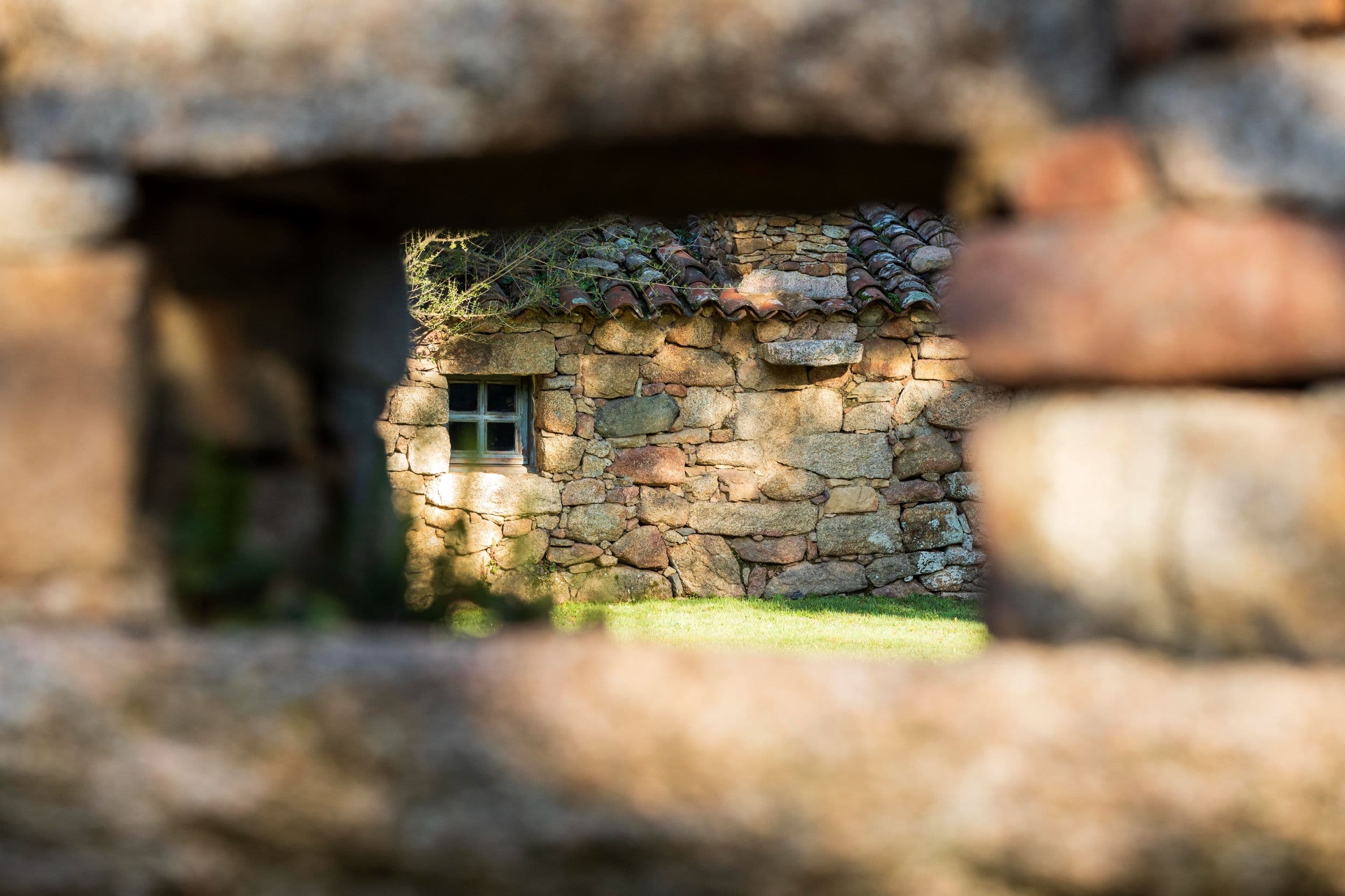 A close detail through a stone gateway — a smaller bergerie roof and a single shuttered window framed inside the pale granite of an outer wall.