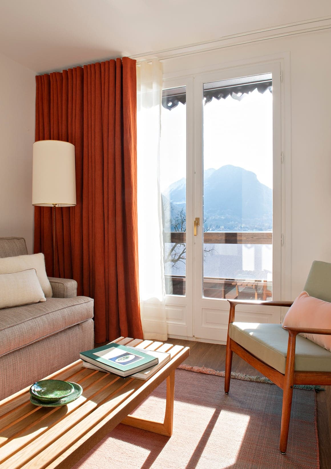 A guest room with terracotta curtains, a mid-century chair set before a French door open onto the lake.
