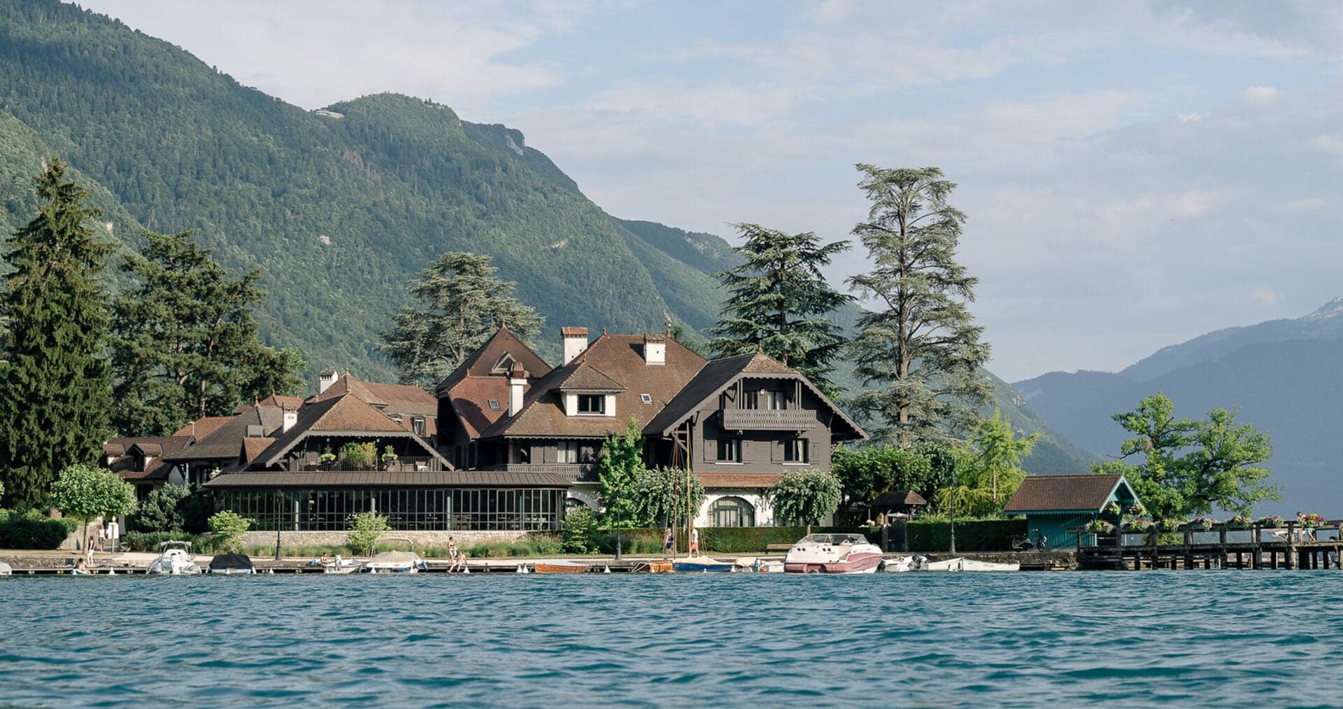 The auberge at Père Bise seen from the water, chalet roofs against the alps and a small dock at the lake's edge.