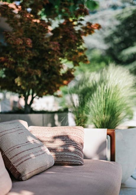 A terrace lounge under a Japanese maple, cushions on a long sofa with the lake just beyond.
