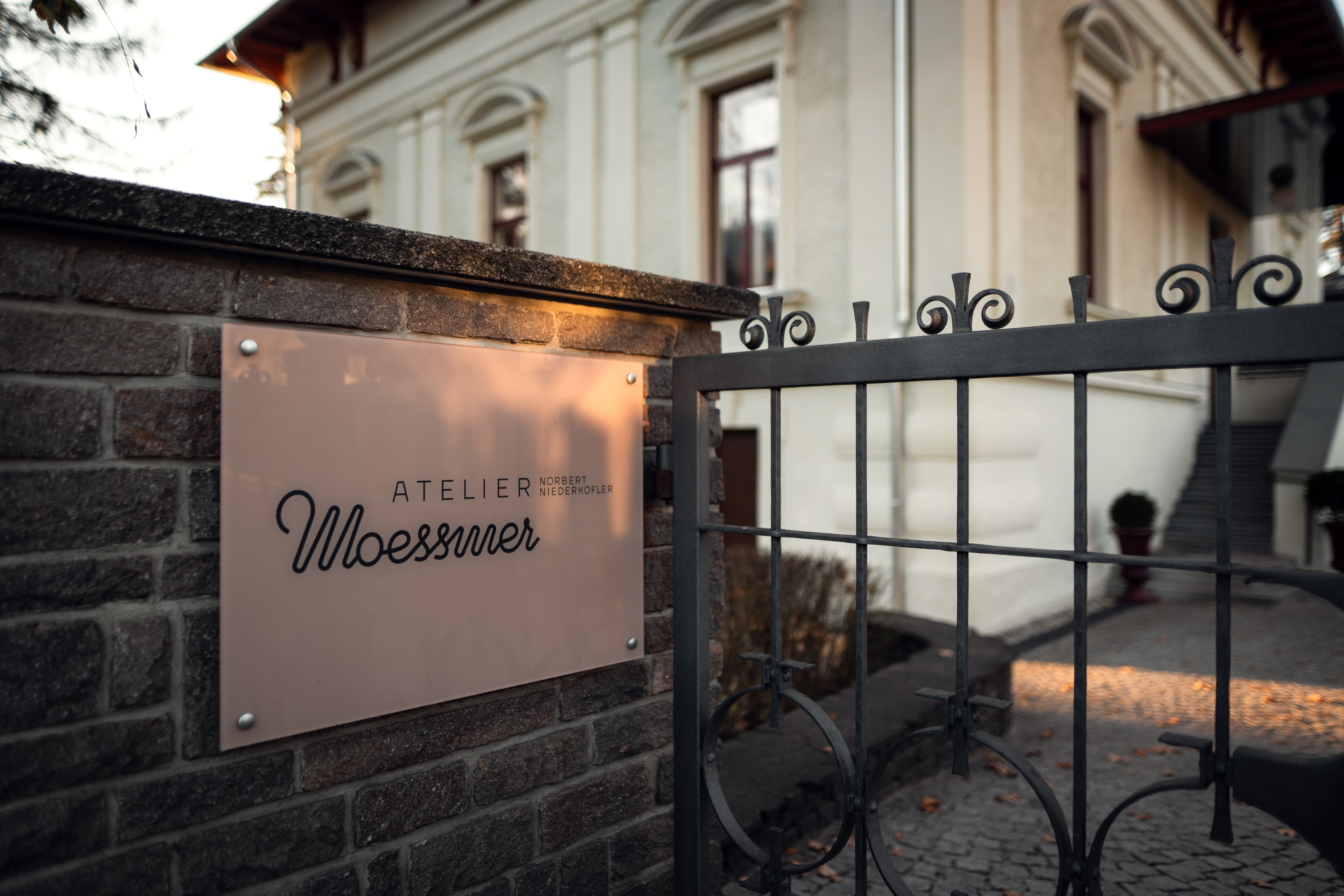 A wrought-iron gate and a stone wall with a small pale-pink plaque reading *Atelier Moessmer Norbert Niederkofler*, the late-classical manor house behind in autumn light.