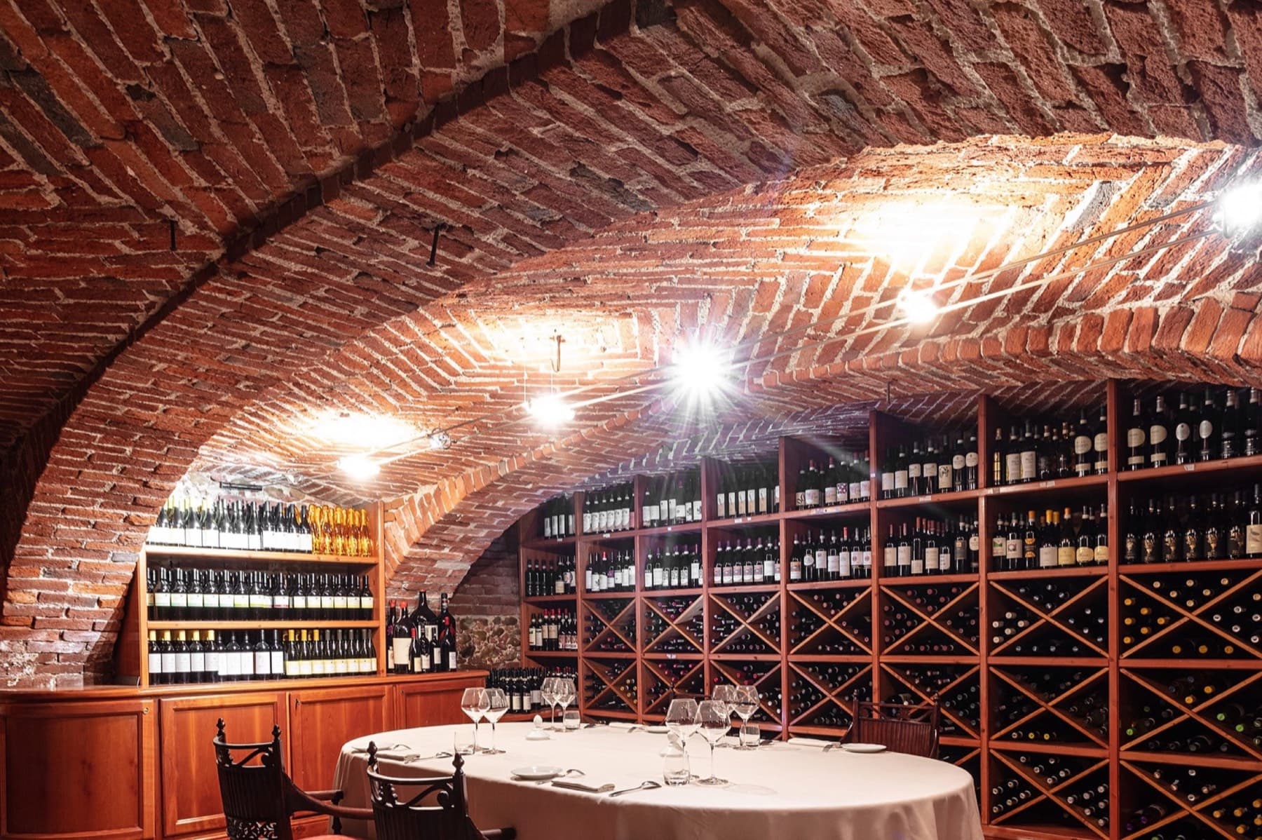 The vaulted brick cantina at Antica Corona Reale — a low Roman-tile arch in warm terracotta, tall walnut wine racks lining both walls, a single round table laid in white linen and crystal beneath the vault.