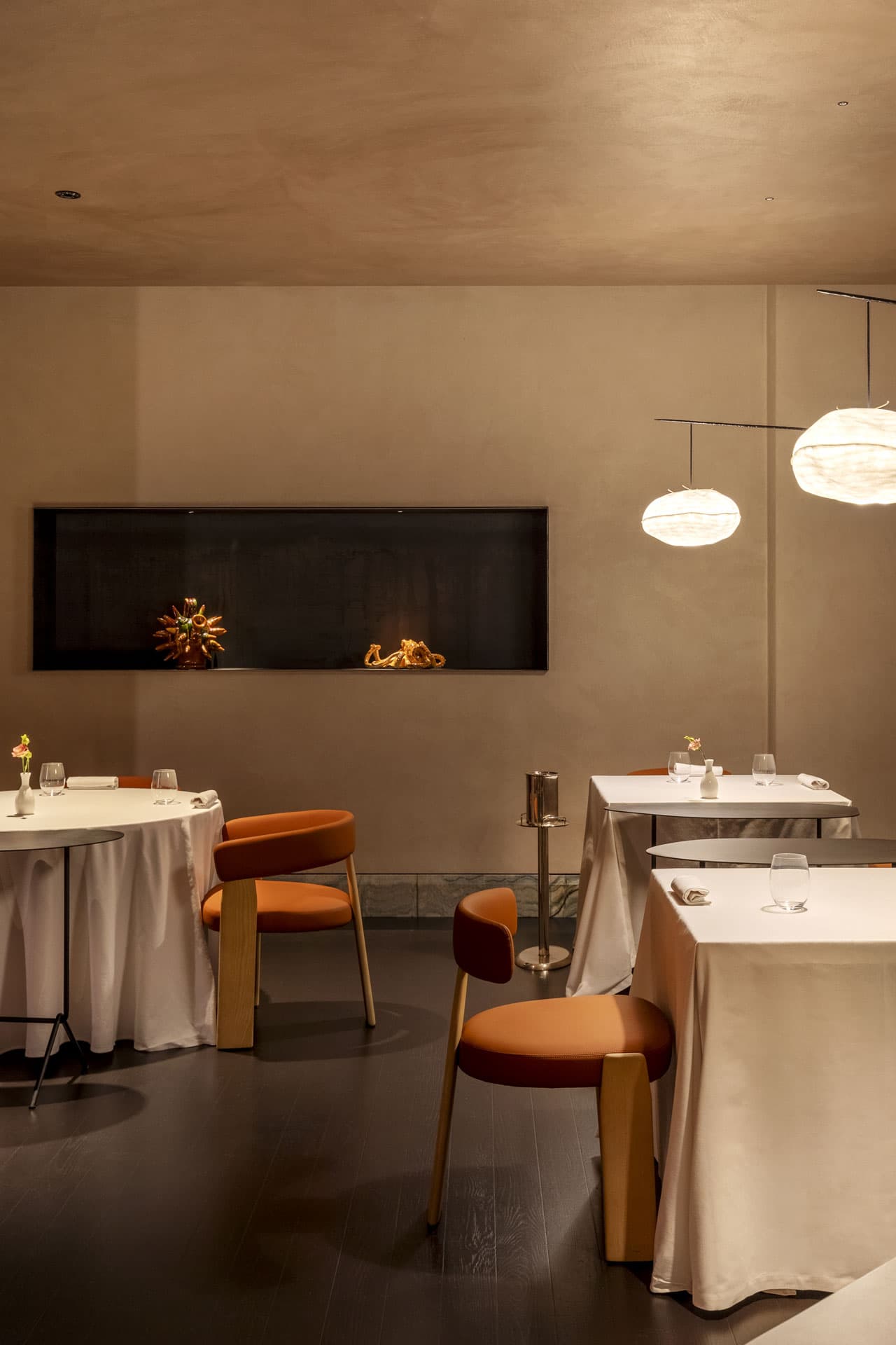 The main dining room at La Madia — soft beige plaster walls, terracotta-leather chairs around white-clothed tables, two paper-cloud pendant lamps, and a long horizontal niche set with a small ceramic octopus and a pinecone vase against burnished black metal.