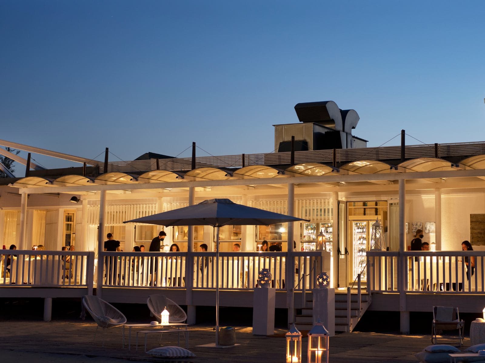 Ristorante Uliassi at dusk — a long, low pavilion of slatted white timber lit from within, lanterns on the terrace, a single white parasol open over the deck and the deep blue Adriatic sky settling above the arched skylights of the roof.
