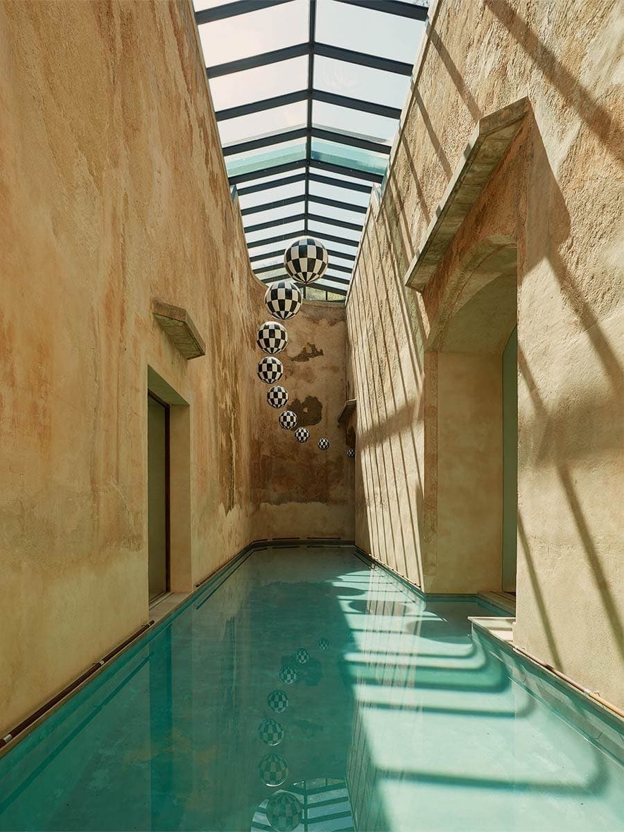 A long, narrow lap pool in the spa, walls of warm sandstone above and a hanging line of black-and-white spheres reflected in the still water.