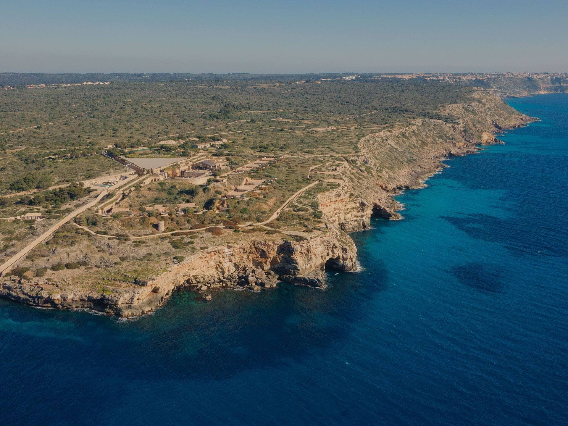 An aerial view of Cap Rocat — the limestone cape running into the Mediterranean, the fortress buildings set back on the headland, deep blue water below.