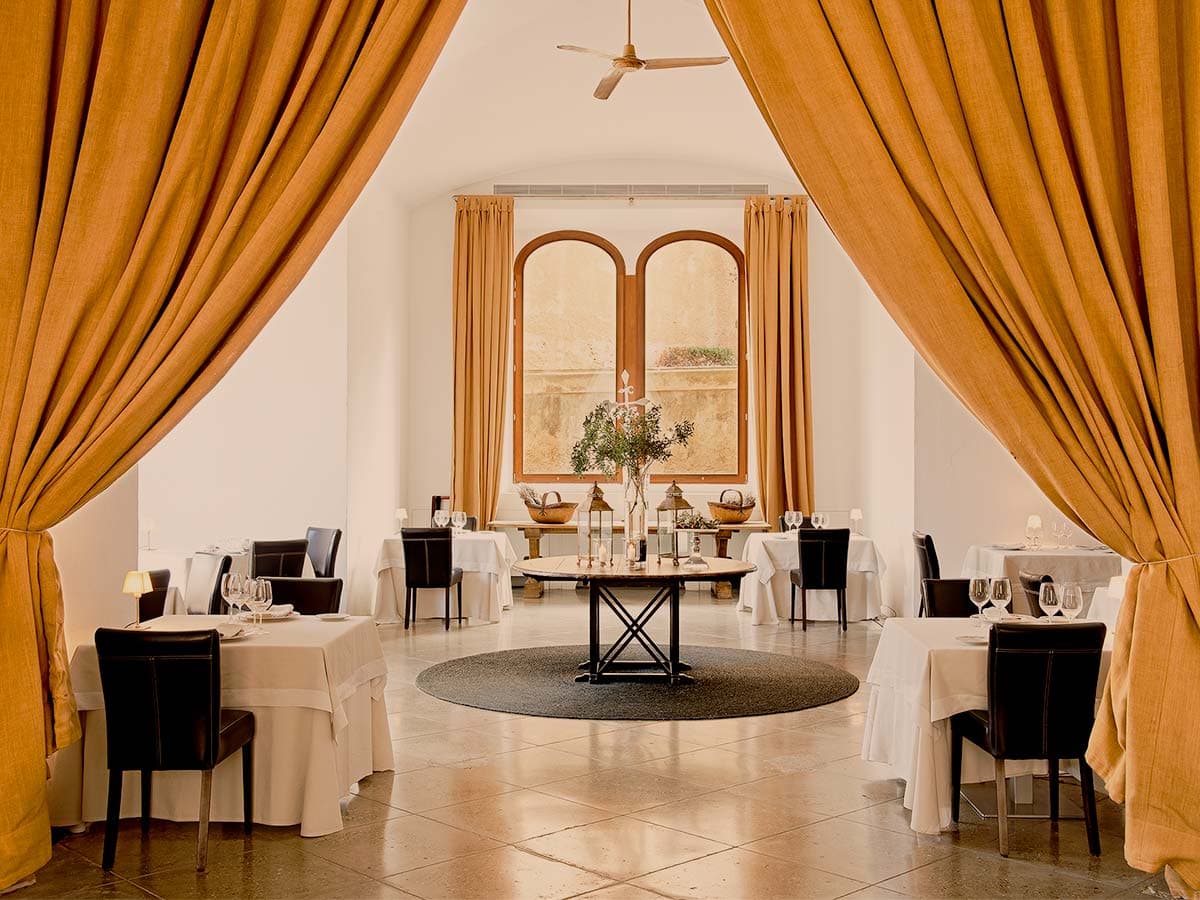 The dining room at La Fortaleza — pale stone floors, white-clothed tables, tall ochre curtains framing arched windows onto the inner courtyard.