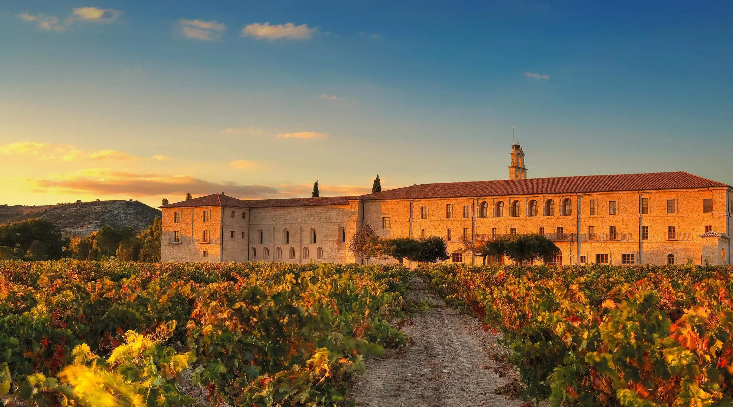 The Abadía Retuerta abbey at sunset, terracotta roofs and a stone bell-tower above autumn vineyards.