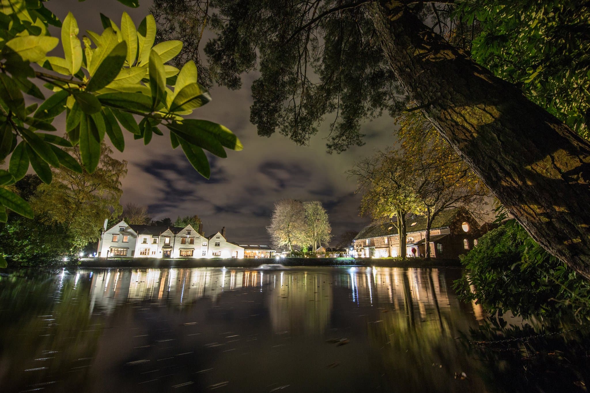 Moor Hall at night seen across its lake — a row of pale-rendered gabled buildings with chimneys and lit ground-floor windows reflected on still water, a low fountain plume catching the lights between bare branches in the foreground.