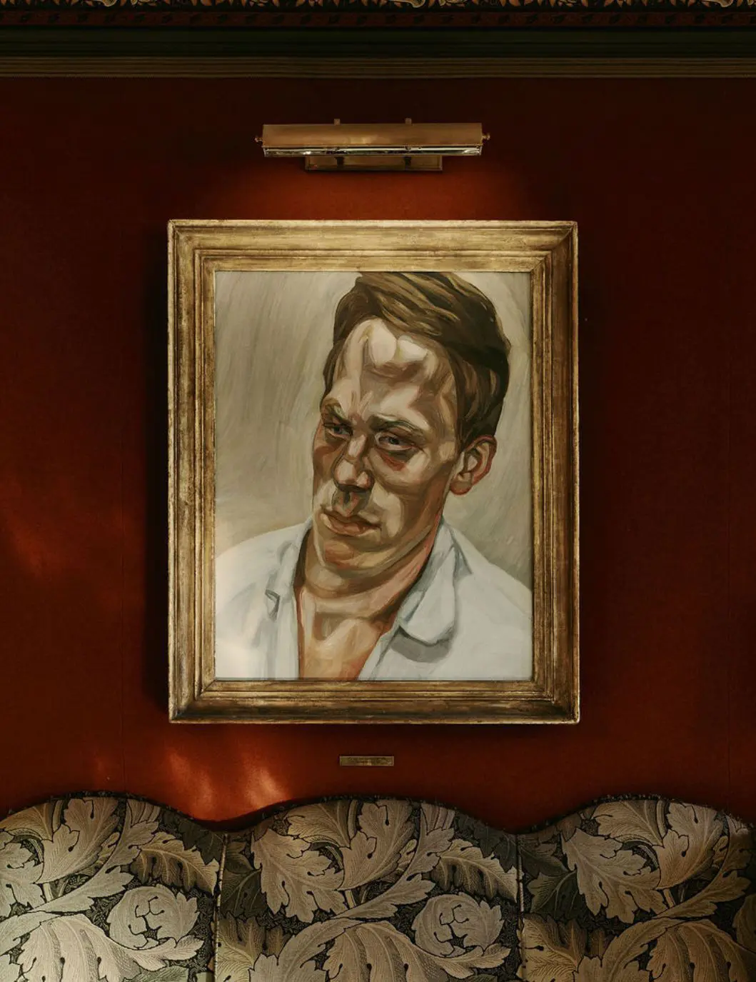 A Lucian Freud portrait hung against a deep red wall, lit by a single picture light above a damask banquette.