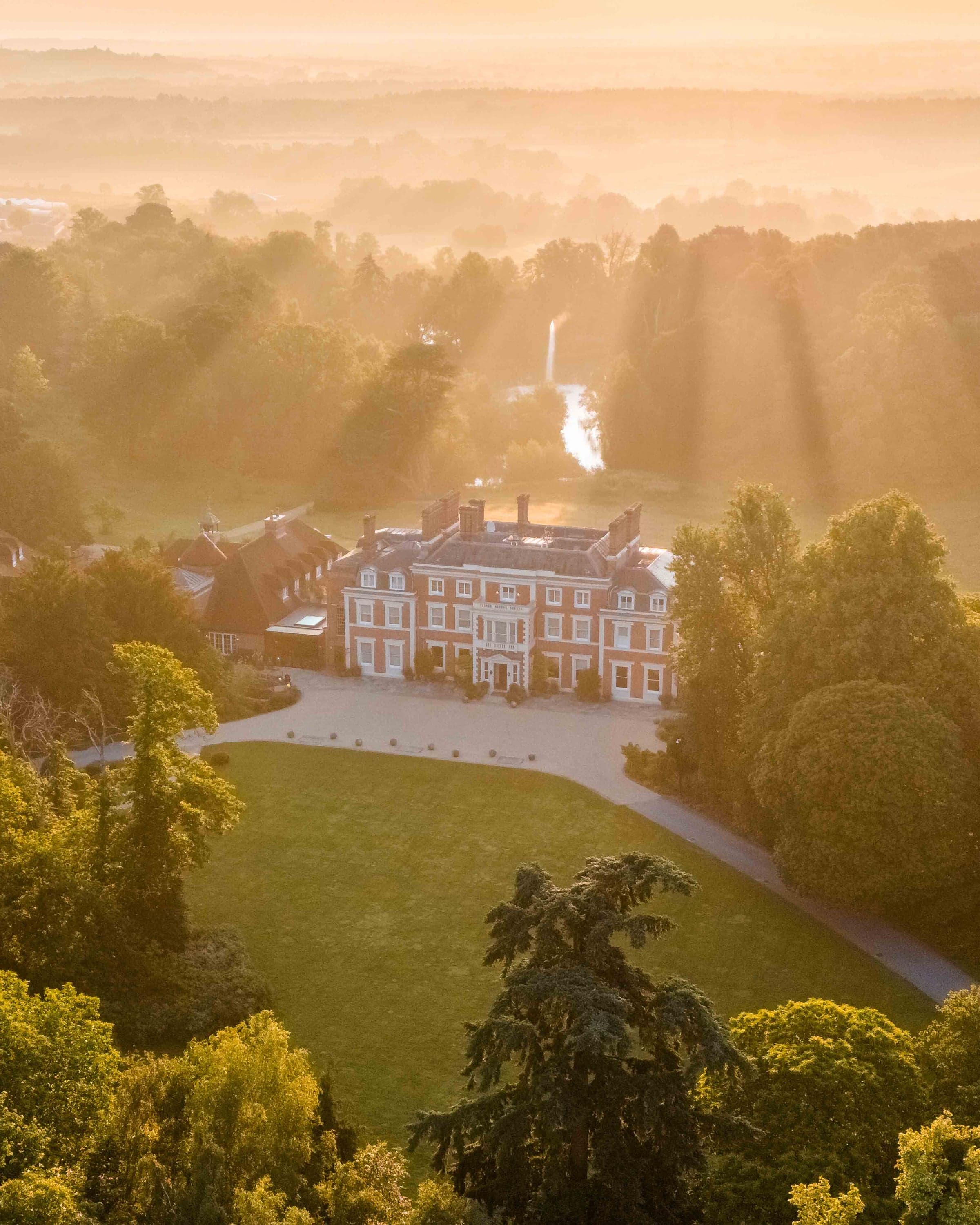A Georgian country house at sunrise from above, sunbeams cutting through the trees that surround it.