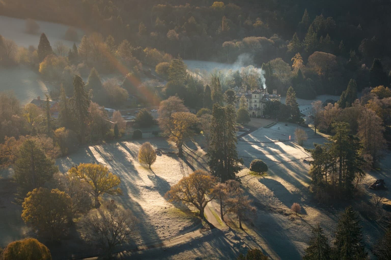 An aerial view of Palé Hall in the Dee Valley at dawn, frost on the lawns and chimney smoke rising into the trees.