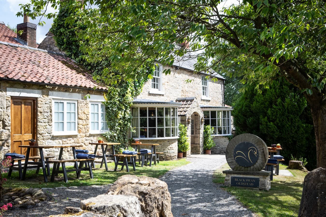 The Black Swan at Oldstead — pale-stone pub with red pantile roof, picnic tables on a gravel terrace, a slate stone carved with the Black Swan emblem, ivy and chestnut leaves catching the summer light.