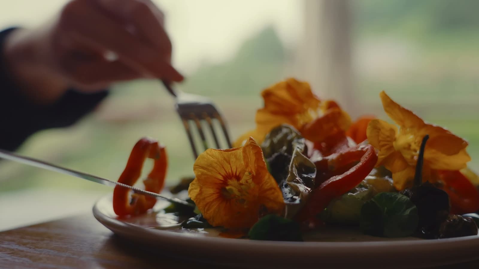 A close view of a plate of leaves, nasturtium flowers and roasted pepper, a hand lifting a forkful in soft window light.