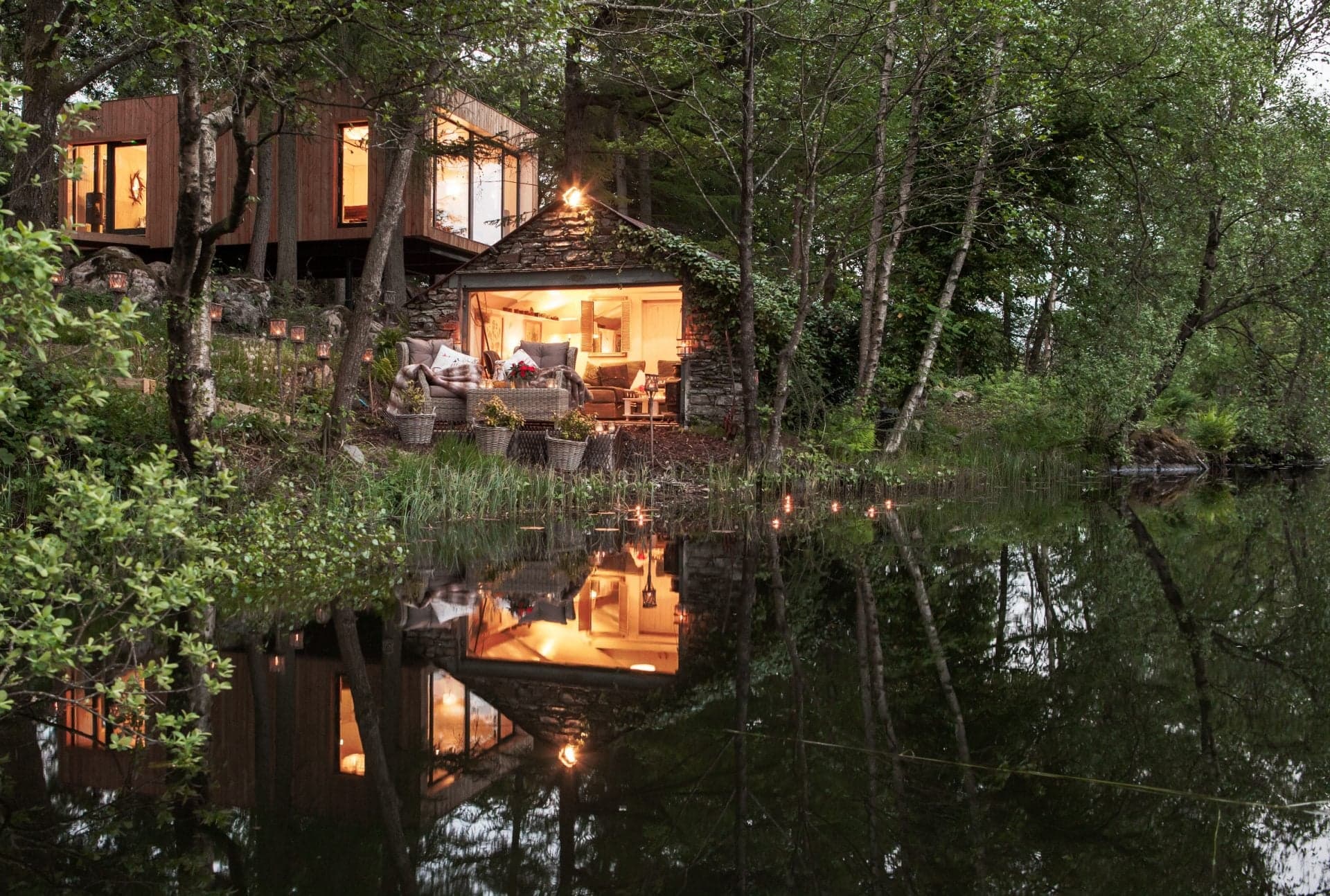 A spa lodge at dusk, lit from within and reflected on the surface of a small pond cut into the woods.