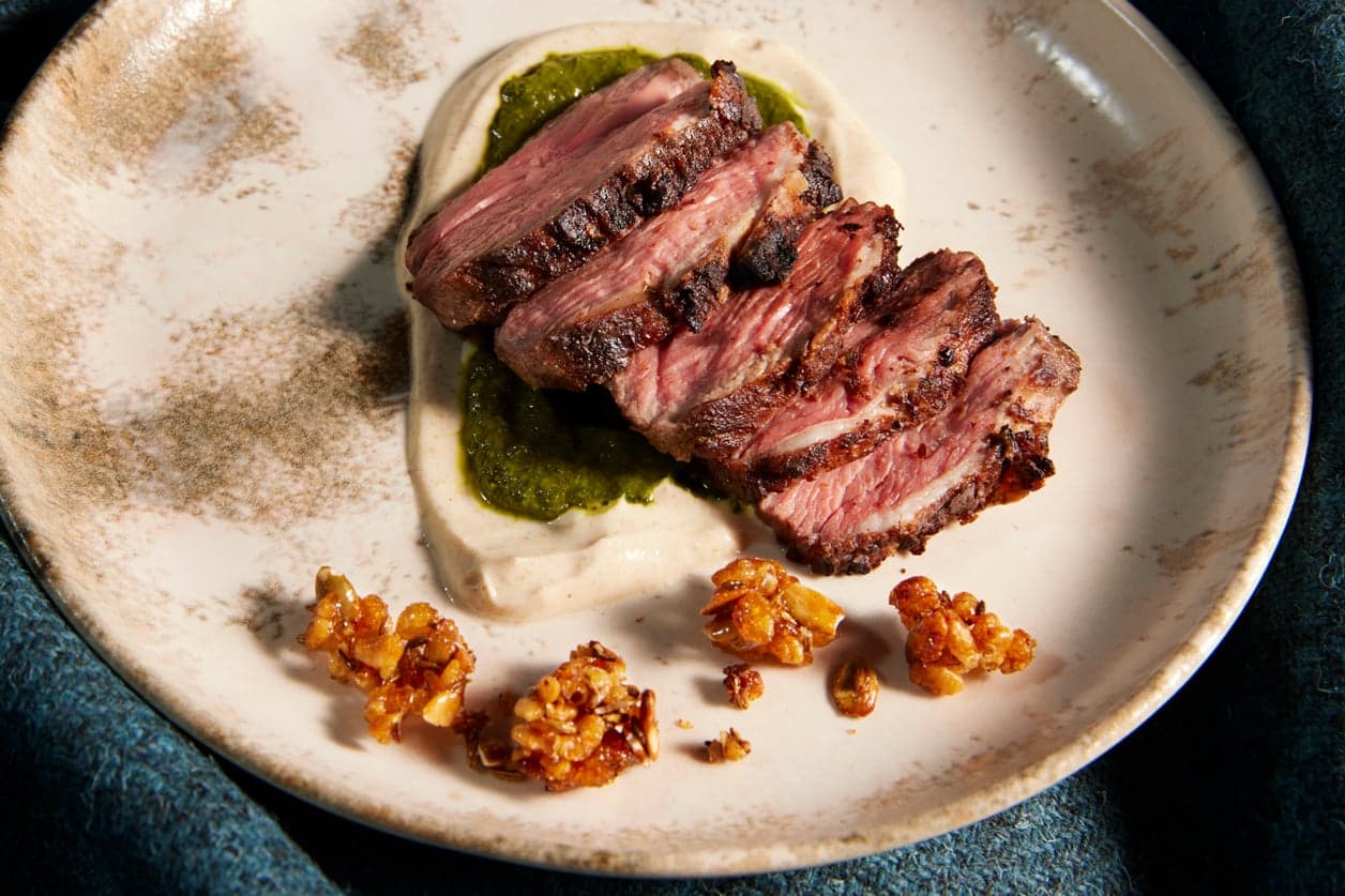 A close-up of sliced lamb resting on a yoghurt smear with a green chutney and crisp puffed-grain garnish.